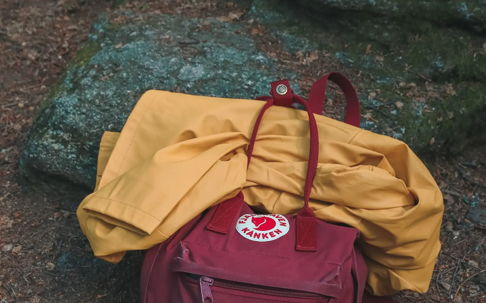 Damp yellow rain jacket draped over a red backpack on mossy ground