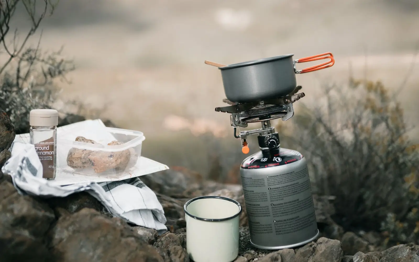 Portable camp stove with a pot boiling beside a mug and food container on a misty hillside